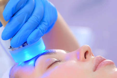 Portrait of woman on apparatus hardware phonophoresis face procedure with hyaluronic acid gel in cosmetology clinic, side view. Cosmetologist beautician doctor moving manipula with cooling blue light.の写真素材