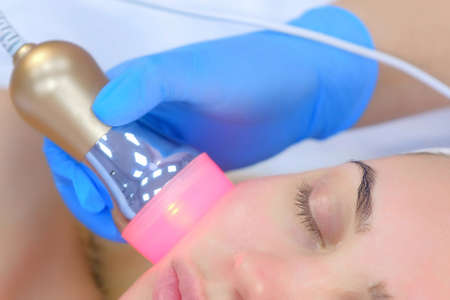 Portrait of woman on apparatus phonophoresis face procedure with hyaluronic acid gel in cosmetology clinic, closeup view. Beautician moving manipula with red light on face. Beauty industry concept.の写真素材