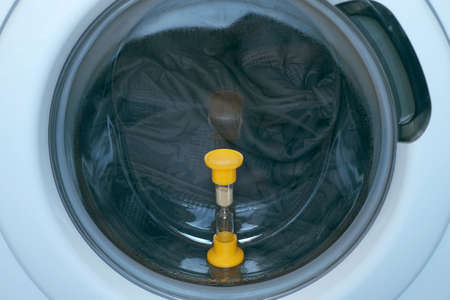 Yellow sandglass on window of washing machine with grey bedspread inside is working, laundry at home. Modern technologies in everyday life, housework and household.の写真素材