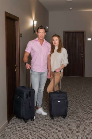 Man and woman with suitcases near their room in hotel. Young family couple looking at camera....