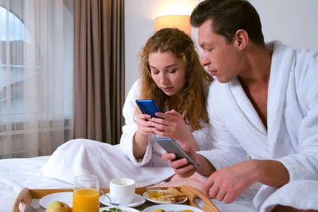 Man and woman are having breakfast in hotel room and reading news in their smartphones talking each...