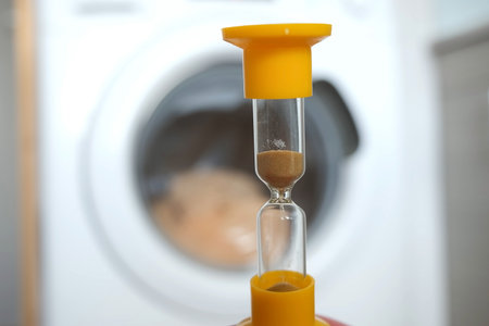 Yellow sandglass on window of washing machine with grey bed linen inside is working, laundry at...
