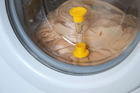 Yellow sandglass on window of washing machine with grey bed linen inside is working, laundry at home. Modern technologies in everyday life, housework and household. Hourglass closeup view.の写真素材