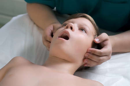 Session of craniosacral therapy, cure of teen boys jaw and neck by a doctor therapist. Craniosacral therapist touches the boys cheeks and checks corrects of the jaws by hands at the hospital.の写真素材