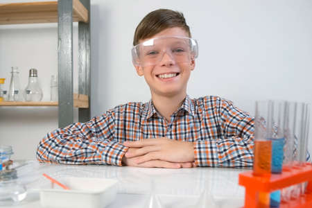 Happy teen boy in protective glasses is sitting at table in chemical laboratory. Online education, homeschooling and science concept. Scientific activity at an early age.の写真素材