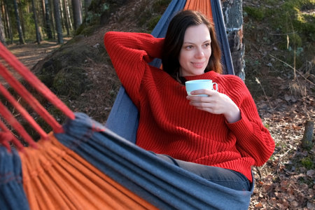 Happy woman relaxing lying in hammock drinking tea in forest admiring nature. Resting relaxed brunette female enjoying time in camping in woodland. Tourist, traveler, camper contemplating outdoors.の写真素材
