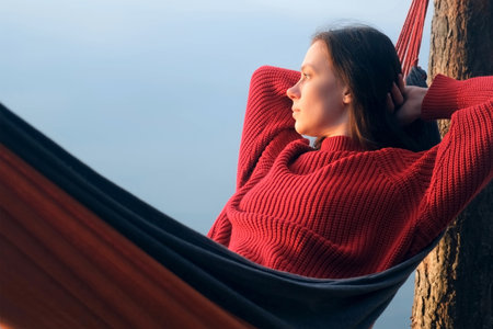Woman relaxing admiring lake lying in hammock in forest at sunset on nature. Resting young female in red sweater enjoying time in camping in woodland. Tourist traveler, camper outdoors.の写真素材