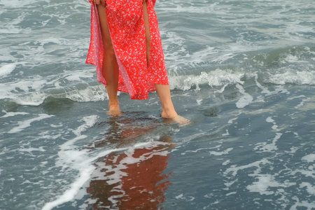 Barefooted young woman wearing red dress walks on wet black volcanic sand enjoying feeling of waves...