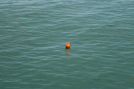 Orange buoy floating on calm water surface with small ripples. Orange maritime safety buoy gently swims on slightly dark turquoise sea, providing navigational guidance across serene oceanic expanse.の写真素材