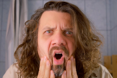 Long haired bearded man shocked by his reflection in mirror in bathroom in morning. Lack of sleep,...