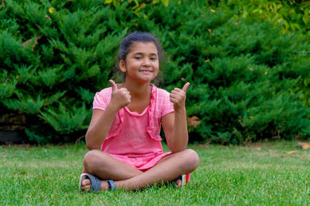 Little child gypsy girl showing thumbs up sitting in grass with little friend text on t-shirtの写真素材