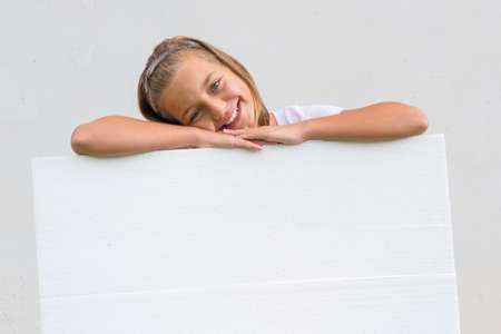 Smiling happy cute child girl lean heands and head over blank banner holging it and presenting as empty sheetの写真素材