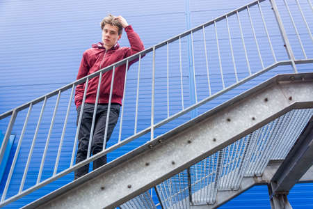 Bored teen boy standing on steel stairs of emergency exit waiting for other person; full length portrait over blue urban building contrast backgroundの写真素材