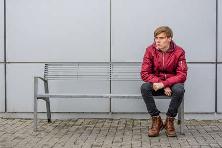 Bored and dissatisfied teenager boy sitting outside of shopping center building in city making faces to express negative thoughtsの写真素材