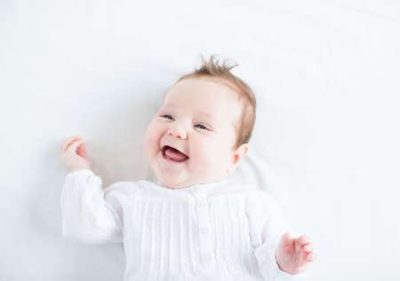 Adorable baby girl in a white dress laughing toothless on a white blanket の写真素材