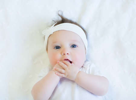 Cute baby girl in white shirt on white blanket with white bow の写真素材