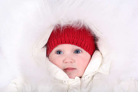 Adorable baby girl looking seriously into the camera wearing a red hat and white fur hood 