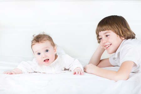 Cute boy and his little sister dressed in white playing on a white bed の写真素材