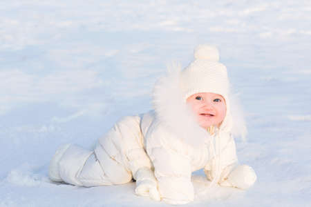 Cute baby in a white fur suit crawling in snow on a very sunny winter day の写真素材