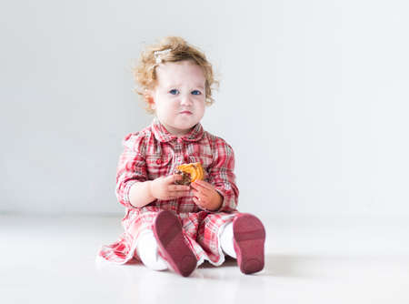 Funny baby girl with curly hair wearing a red dress eating a Christmas cookie in a white nursery の写真素材