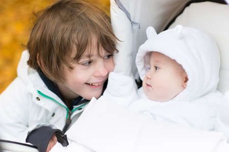 Brother playing with his baby sister sitting in a stroller on a walk in an autumn park の写真素材