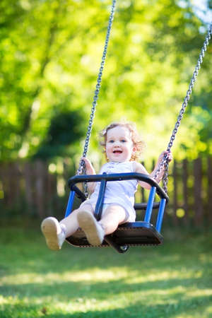 Adorable baby girl with big beautiful eyes and curly hair having fun on a swing ride at a playground in a sunny summer park の写真素材
