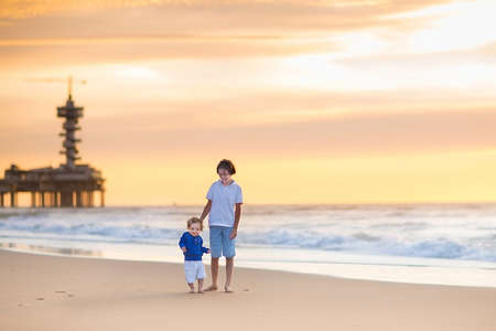 Young brother and his baby sister having fun together playing at a beautiful beach in Holland at sunset on a warm summer evening の写真素材