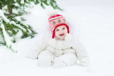 Beautiful laughing baby girl sitting under a Christmas tree wearing a white snow jacket and warm knitted hat の写真素材