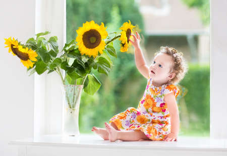 Happy smiling baby girl playing with beautiful sunflowers on a sunny summer day sitting at a window to the garden の写真素材