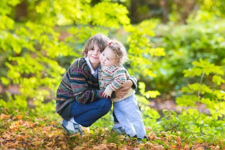 Cute brother and his baby sister hugging and playing together in a beautiful sunny autumn park の写真素材