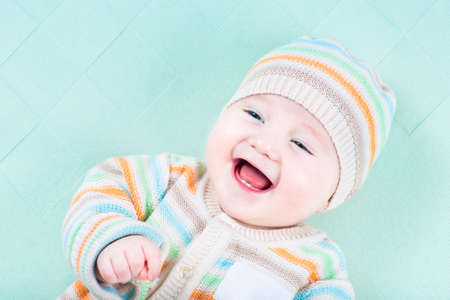 Adorable laughing baby wearing a warm knitted jacket and hat relaxing on a green blanket の写真素材