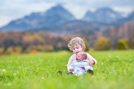 Cute curly toddler girl and her newborn baby brother playing in a field with snow covered mountainsの写真素材