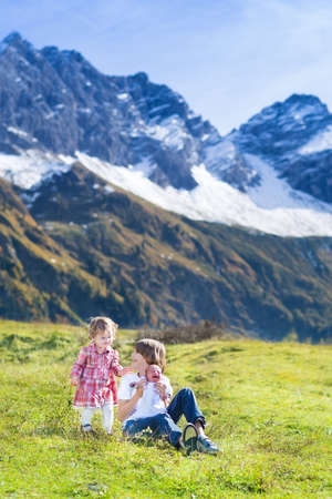 Happy three kids, teenager boy, toddler girl and their newborn baby brother playing in a field between snow covered mountains の写真素材