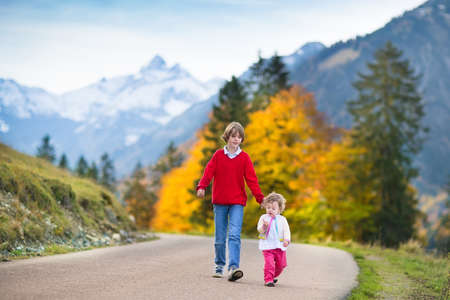 Happy smiling boy and his little baby sister walking on a road between snow covered mountains and yellow autumn trees の写真素材