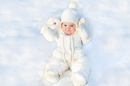 Funny little baby sitting in fresh snow wearing a white jacket and a warm knitted hat の写真素材