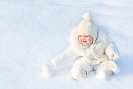 Beautiful little baby girl sitting in white snow wearing a warm jacket and knitted hat の写真素材