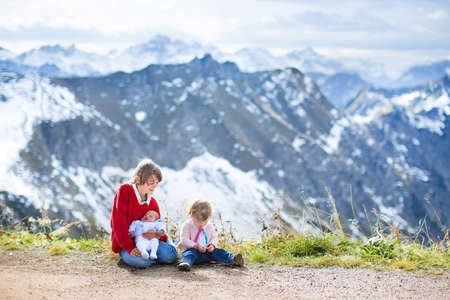 Three children, a teenager boy, toddler girl and their newborn baby brother, playing together between beautiful snow covered mountains の写真素材