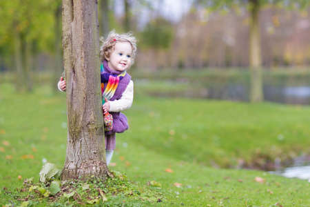 Sweet funny toddler girl hiding behind a tree in a beautiful autumn park の写真素材