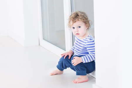 Cute toddler girl sitting at a big window in a white living room 