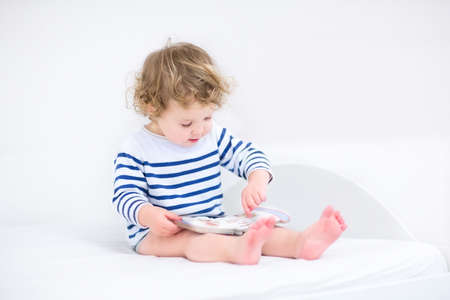 Cute toddler girl reading a book sitting in a white bedroom の写真素材