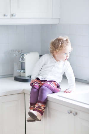 Adorable toddler girl sitting in a white kitchen on the counter top の写真素材