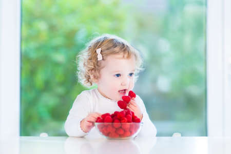 Adorable baby girl eating raspberry at a white table next to a big window の写真素材