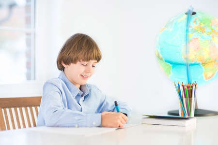 Happy school boy doing his homework at a white desk 