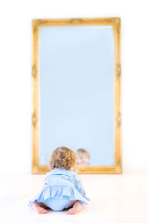 Funny toddler girl with beautiful curly hair wearing a blue dress watching her reflection in a mirror の写真素材