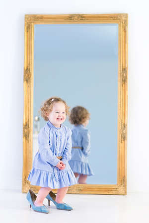 Funny laughing toddler girl with beautiful curly hair wearing a blue dress is trying on her mother s elegant high heels shoes standing in front of a big mirror with wooden frame in a white bedroom の写真素材