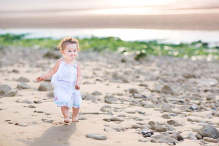 Funny toddler girl running at the beach at sunset wearing a white dress の写真素材