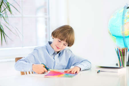 Happy smiling boy cutting colorful paper with scissors in a white desk の写真素材