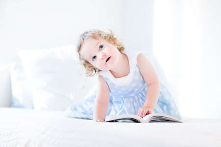 Beautiful little toddler girl with curly hair in a blue dress reading a book on a white bed の写真素材