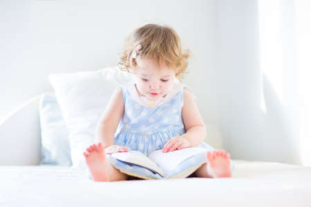 Funny toddler girl in a blue dress reading a book on a white bed in a sunny bedroom の写真素材