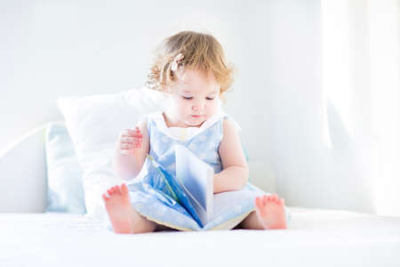 Cute toddler girl with curly hair wearing a blue dress sitting on a white bed in a sunny bedroom reading a book の写真素材